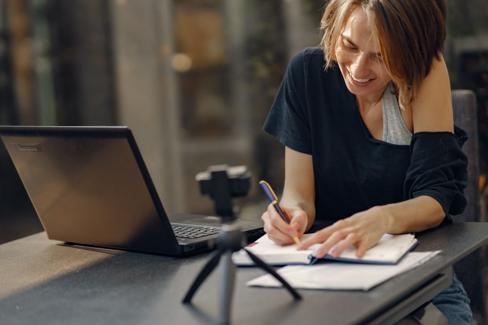 Es zeigt eine Frau, die vor ihrem Computer mit Webcam sitzt und etwas in ihr Notizbuch hinein schreibt.